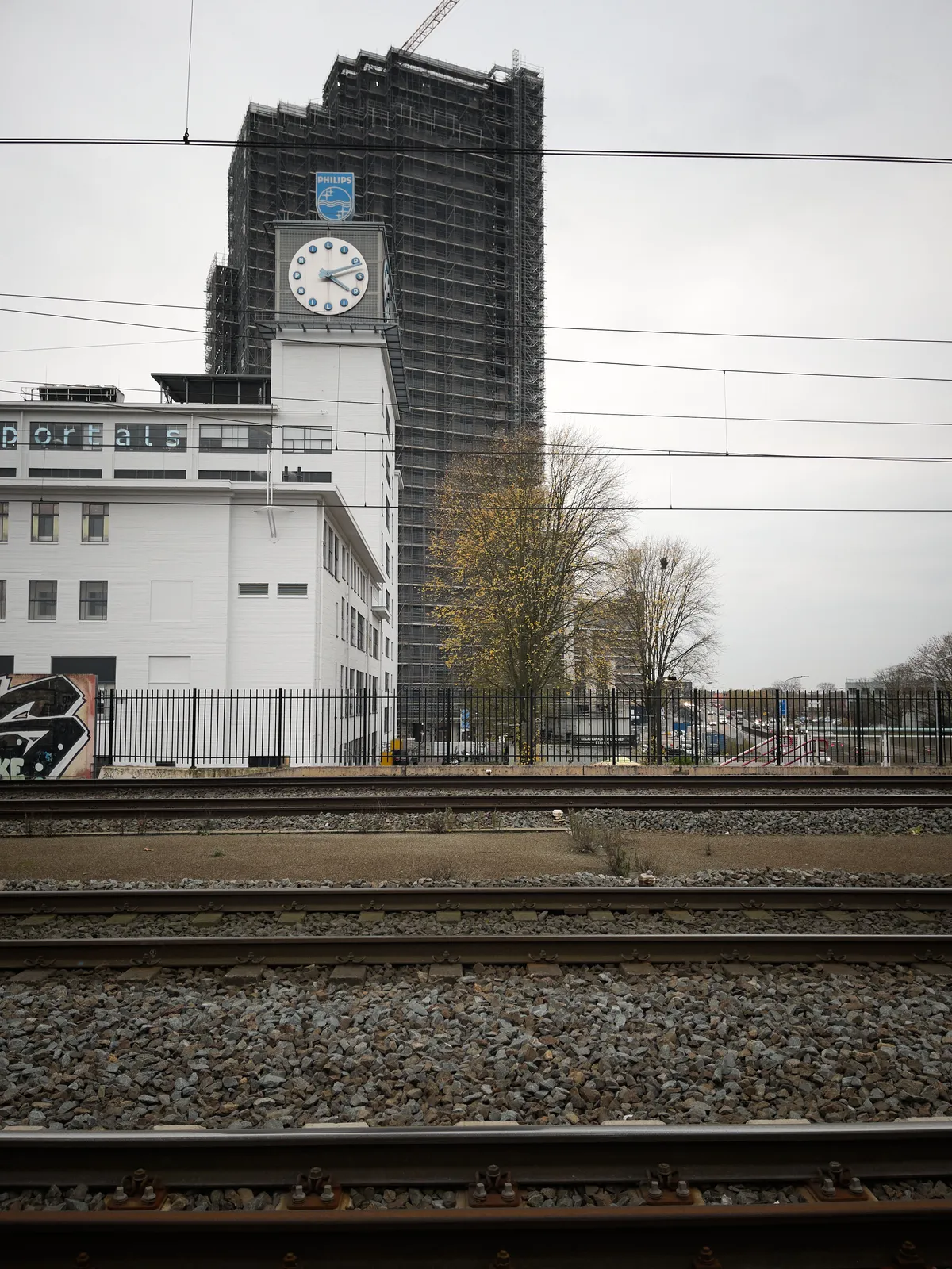 A huge grey building is being built in the background. In front of it is a monumental white building with a very large clock on it. This clock points at 11 minutes past 4.