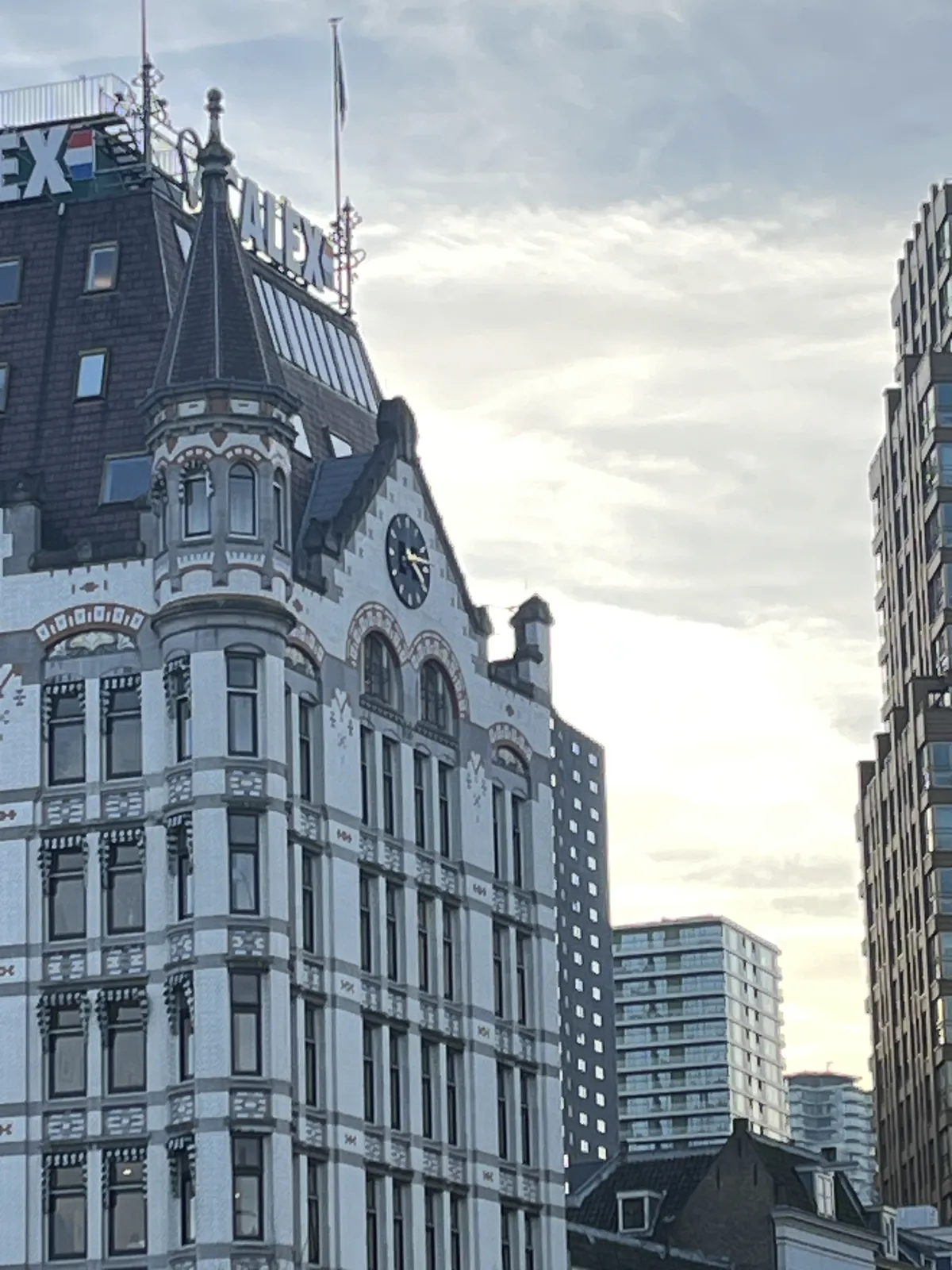A picture of the famous white house in Rotterdam, which some highrise buildings in the background. The clock on the facade of the white house points at approximately 14 minutes past four.