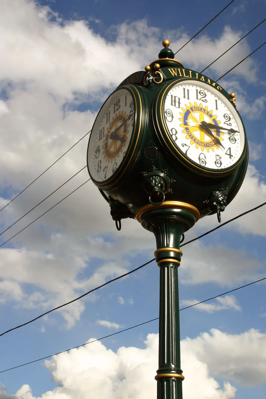A dark green cube on a pole, with seemingly four clocks on it. It is 16:15 according to the two visible sides.