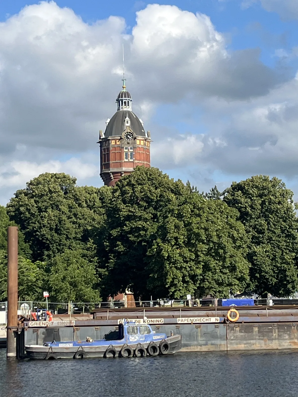 An old watertower towers over some large, green trees. In the foreground is a tugboat, attached to a barge. There’s a clock on the watertower, and it points at approximately 20 minutes past four.