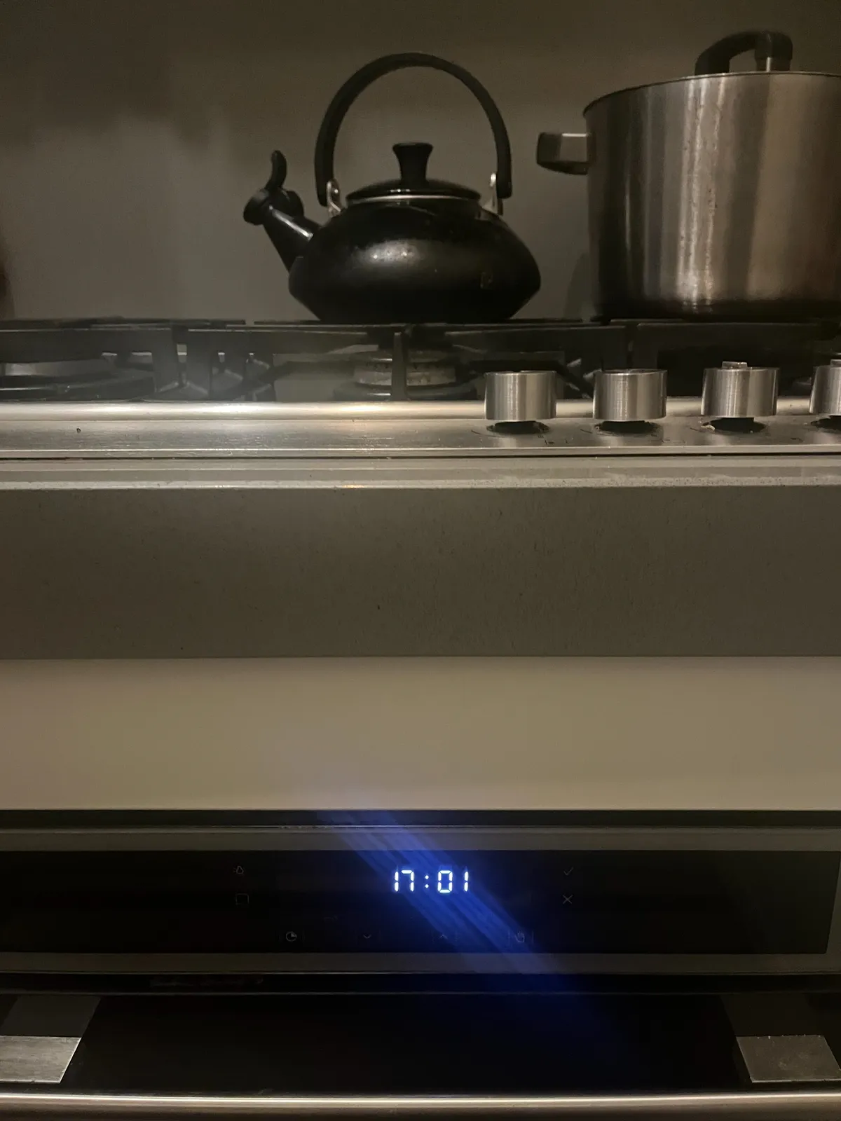 A kettle and a pan on a stove. The stove’s digital clock says it is one minute past 17.