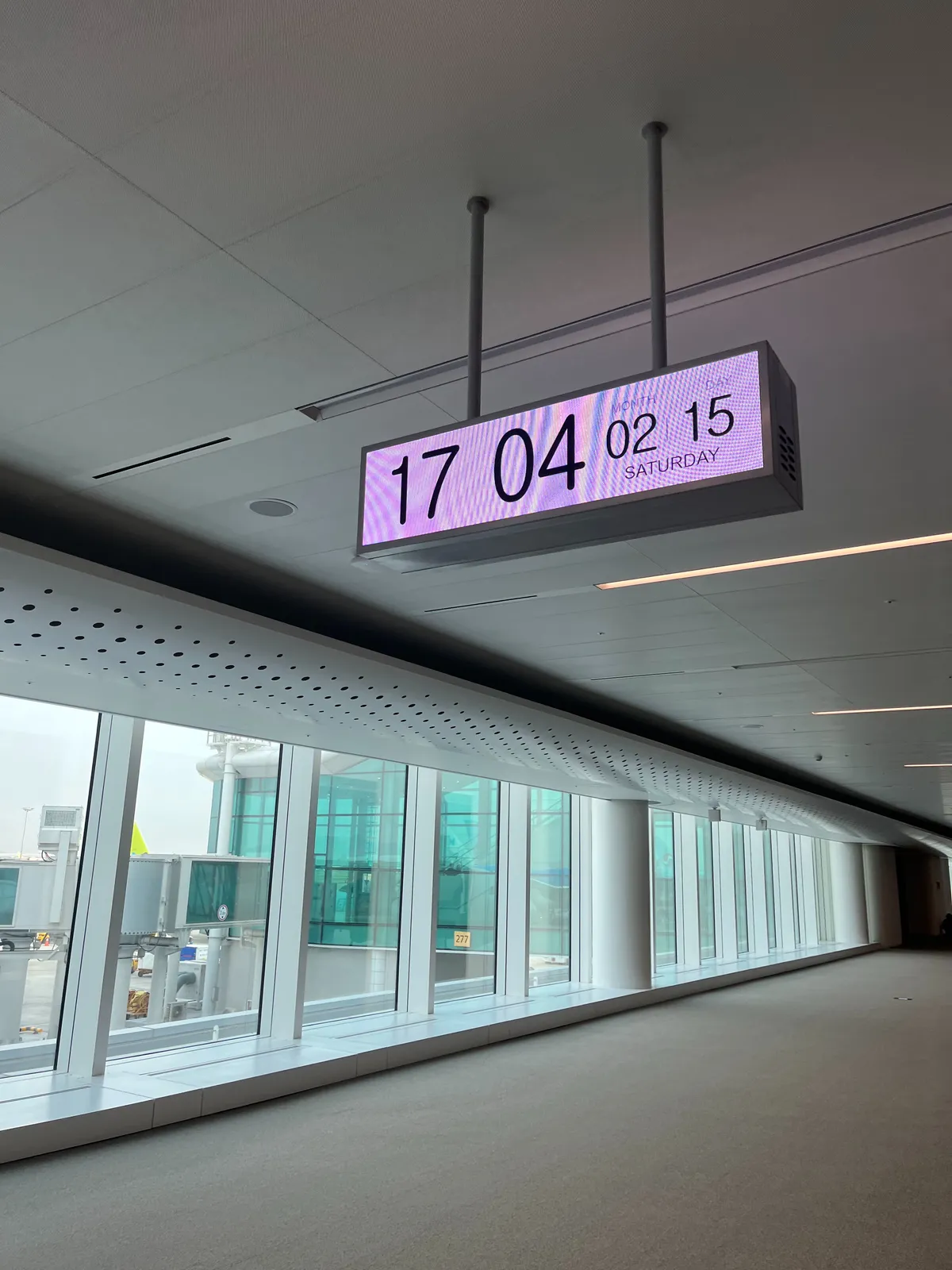 A lit digital clock, in a lit hallway. It says the date is Saturday 15 February, which is almost certainly not true. The time might be right though: it is displayed as 17:04.