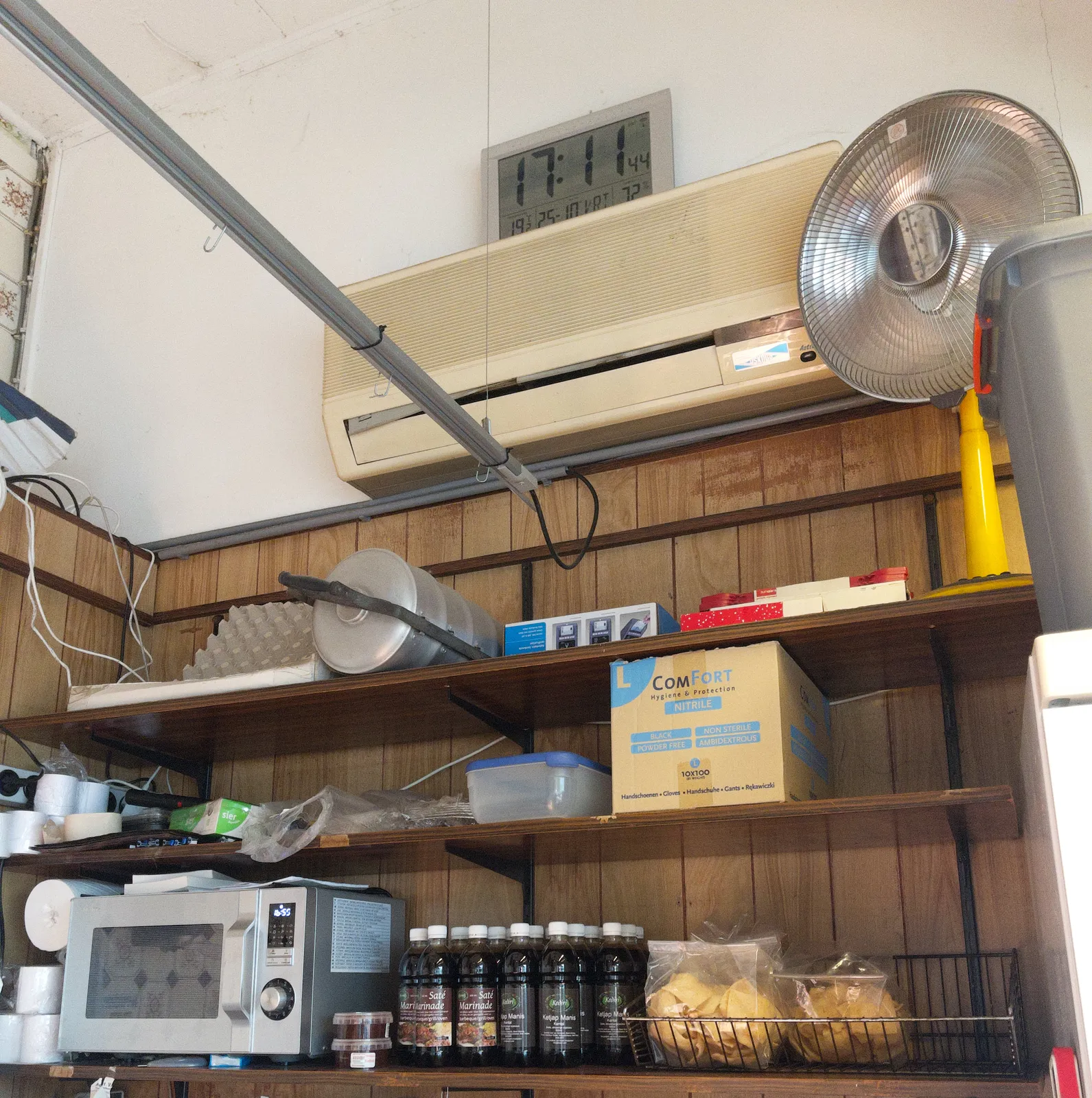 A cupboard with a microwave, some indonesian snacks and pots, a ventilator, an airco, and a large digital clock which says it is 17:11.
