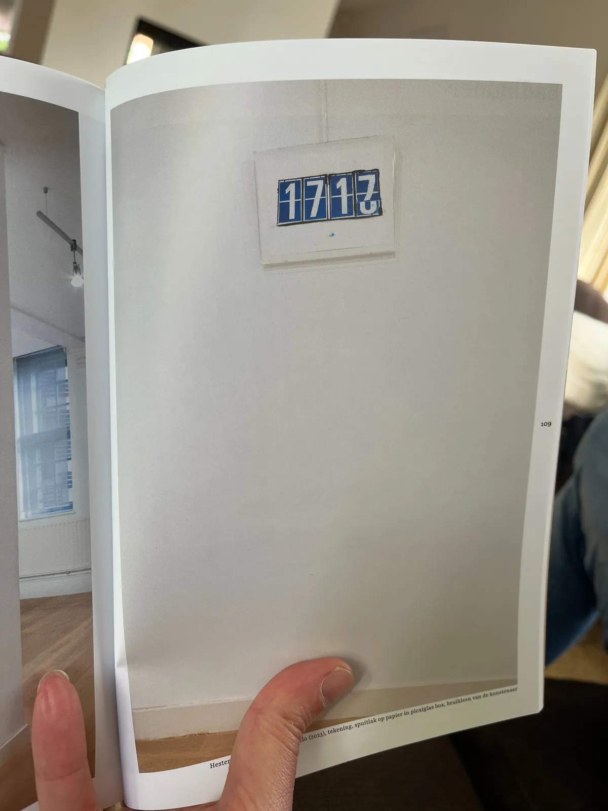 Someone holding a book. On the open page there’s an image of a painting of a flip clock. It is painted in the exact moment that it flips from 17 past 17, to 18 minutes past 17.