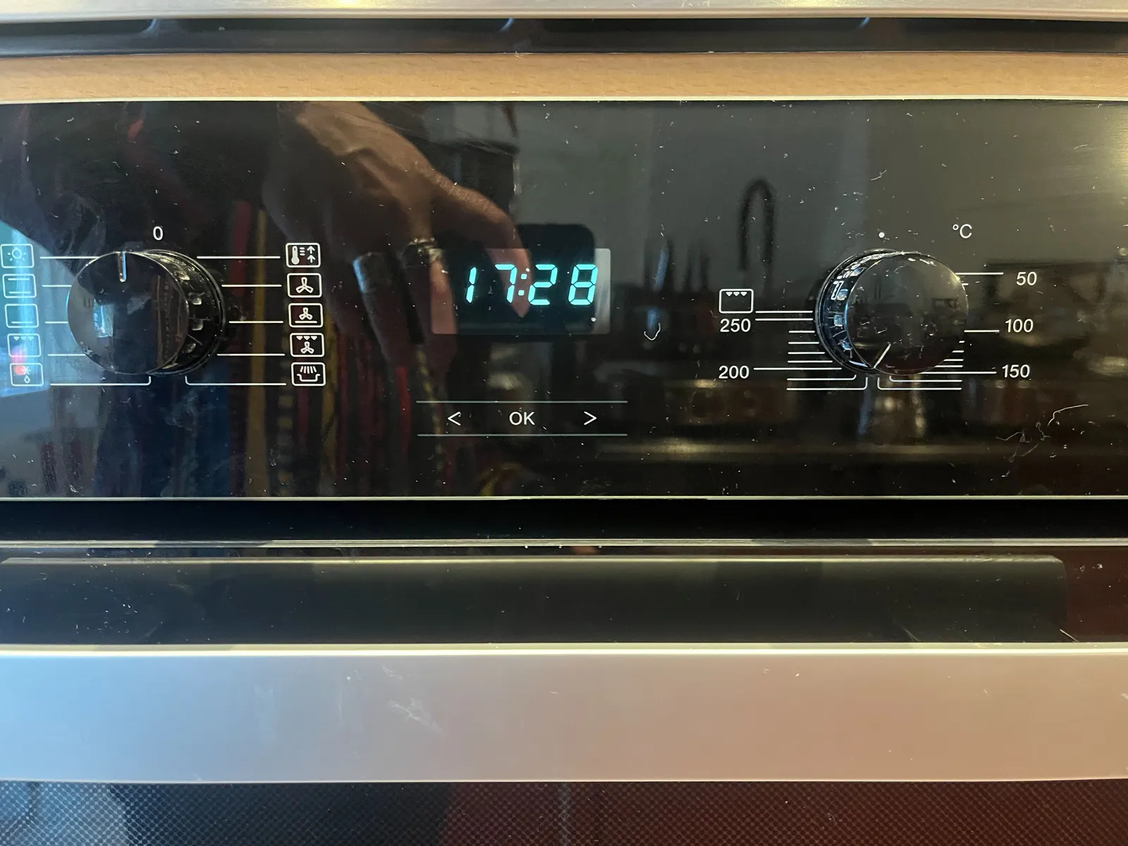 Two knobs of an oven with all kinds of cryptic symbols around them. Between the knobs there’s a digital display that shows the time as 17:28.
