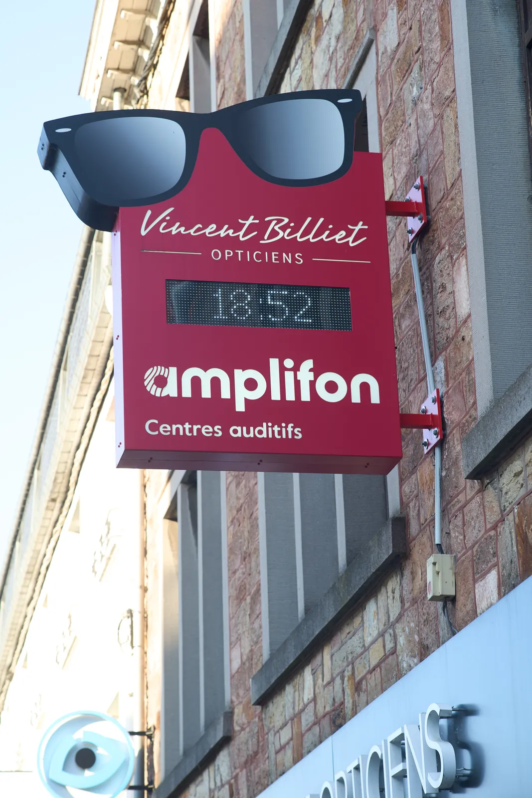 A shop sign for Vincent Billiet opticians. The sign has an icon of large black glasses on it, and it says Amplifon, centre auditifs. The sign also has a very low resolution display that says it is 18:52