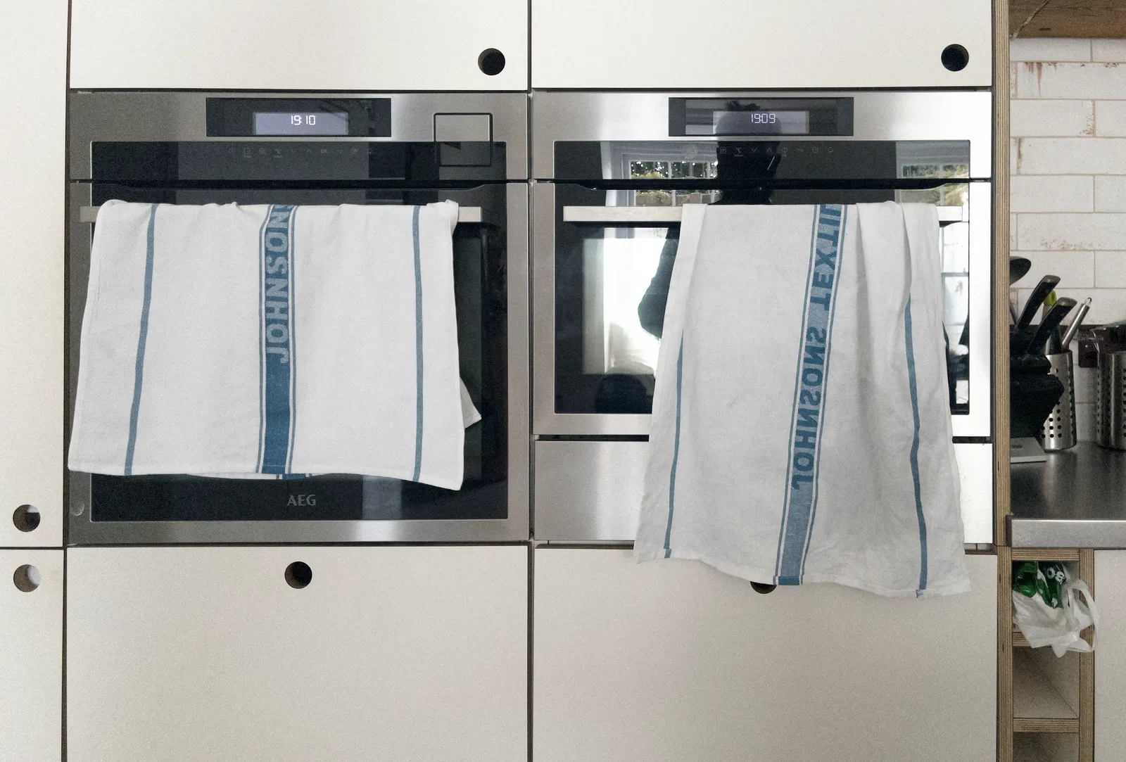 Two identical looking ovens built into a kitchen wall. They both have tea towels draped over them, and they both have a digital display on them that shows the time. Both show a different time though. One of them says it is 19:09, the other says it is 19:10