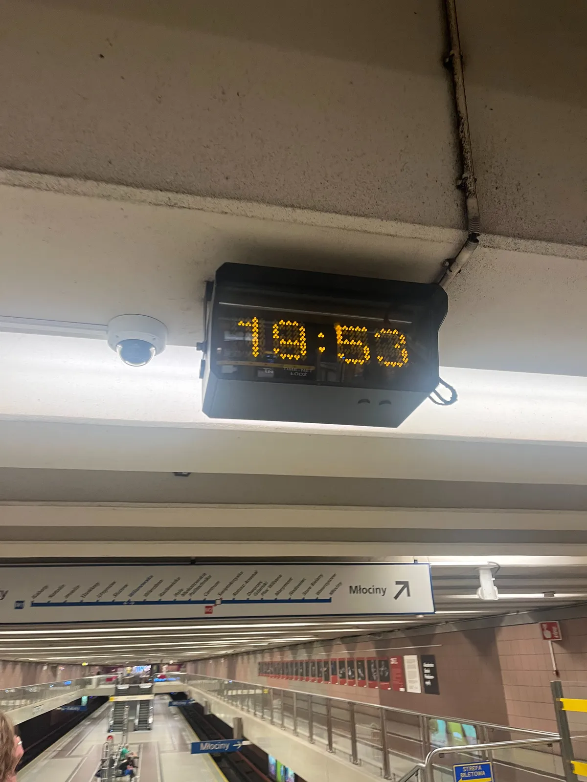 A digital clock attached to the ceiling of a metro station. It says it is 19:53.