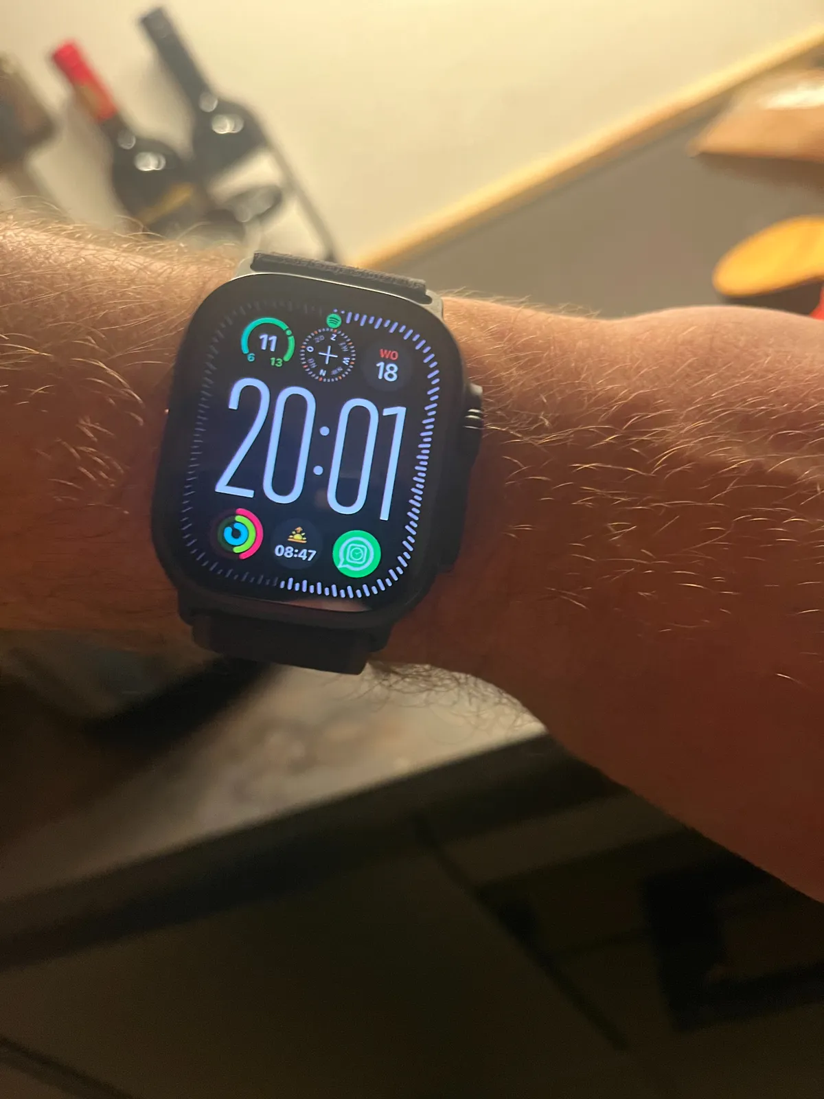 A very large digital watch with all kinds of indicators surrounding the clock. In the background there are two bottles of wine. The watch says it is 20:01