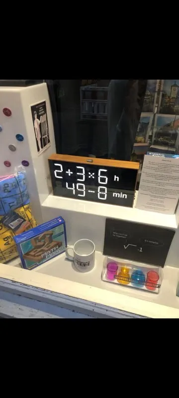 A mathematical clock in a shopping window. It displays the time in two lines of mathematical calculations. The first line says 2 + 3 × 6 h, and the second says 49 - 8 min. Let me repeat that for you, because I had to look twice to understand the time. The first line says 2 + 3 × 6 h, and the second says 49 - 8 min. 