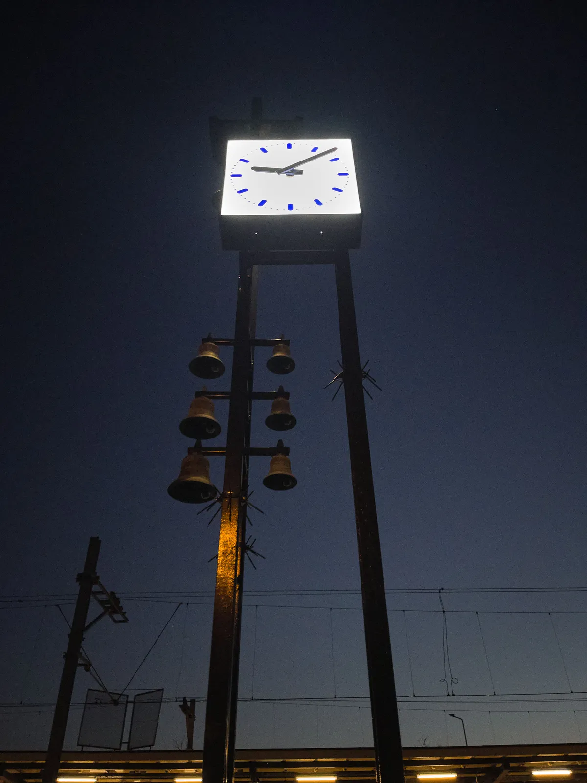 A Dutch station clock high on some metal poles, against a dark sky. Attached to these poles are some bells as well. The clock points at 7 minutes past 9.
