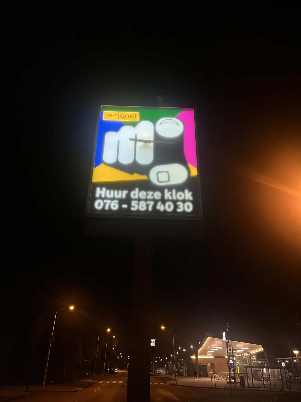 A brightly lit, colourful advertisement on a pole, in a very dark night. In the background, at the bottom of the picture, there’s a deserted train station and a few lamp posts. The advertisement, which is also a clock, is of an abstracted hand which points right at the viewer. The message below this hand says you can rent this clock, with a phone number printed below it: 0765874030. The clock itself points at sixteen minutes past nine.
