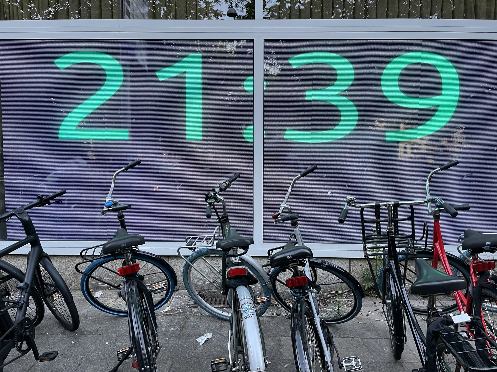 A series of parked bicycles in front of a wall which is covered in LED lights, which show the time as 21:39