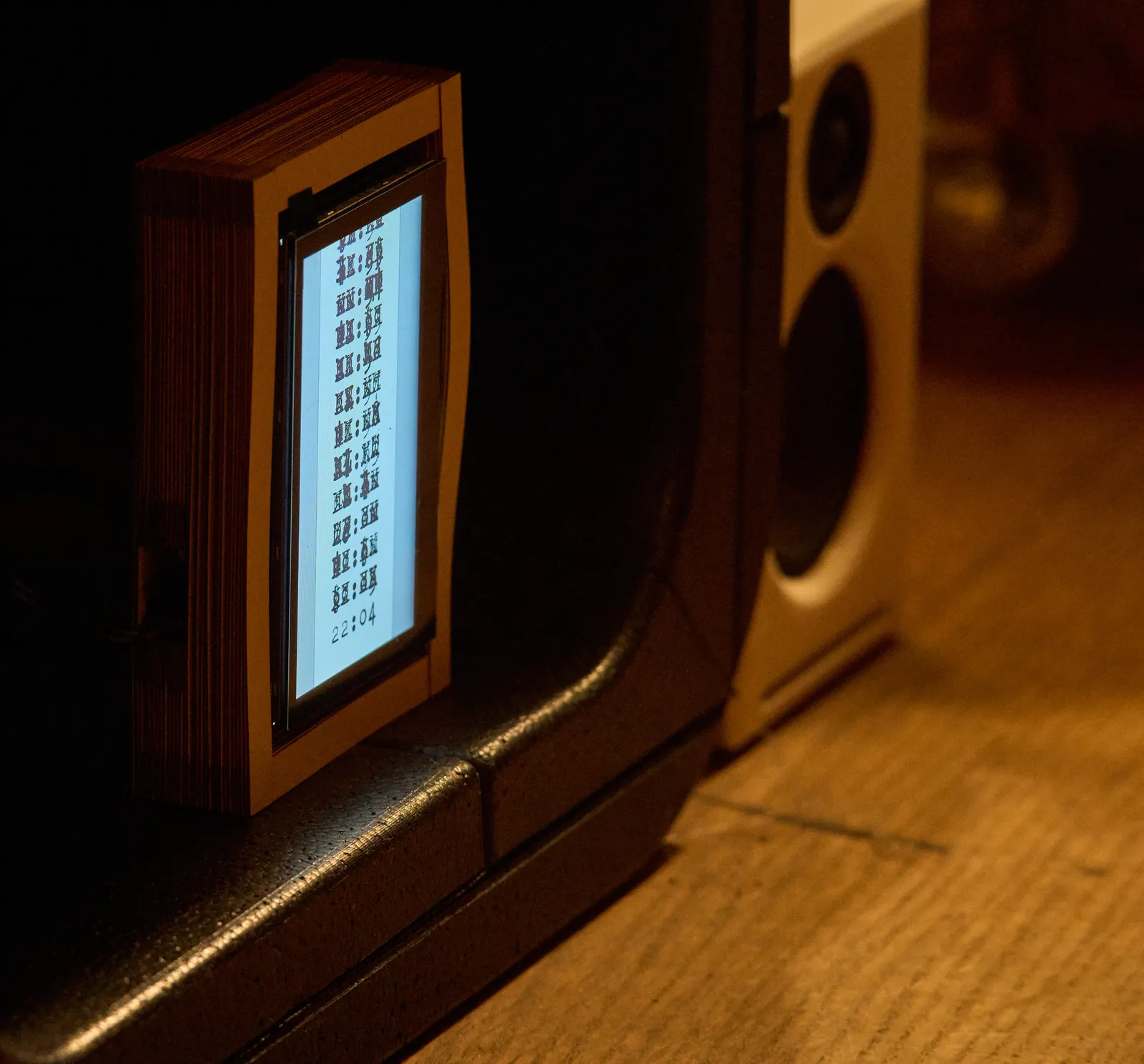 An arty clock on some sort of cupboard. Behind it there’s a little speaker. On the screen of the arty clock you can see a few lines that look like they were typed over a few times by a typewriter. Only the lower line is readable. It spells 22:04.