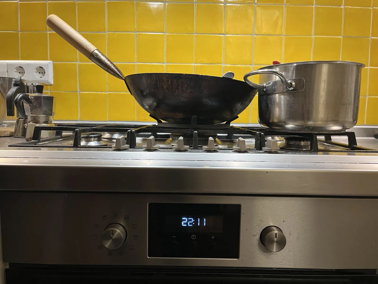 Two pans on a stove, with yellow green tiles behind them. The clock on the stove displays the time as 22:11