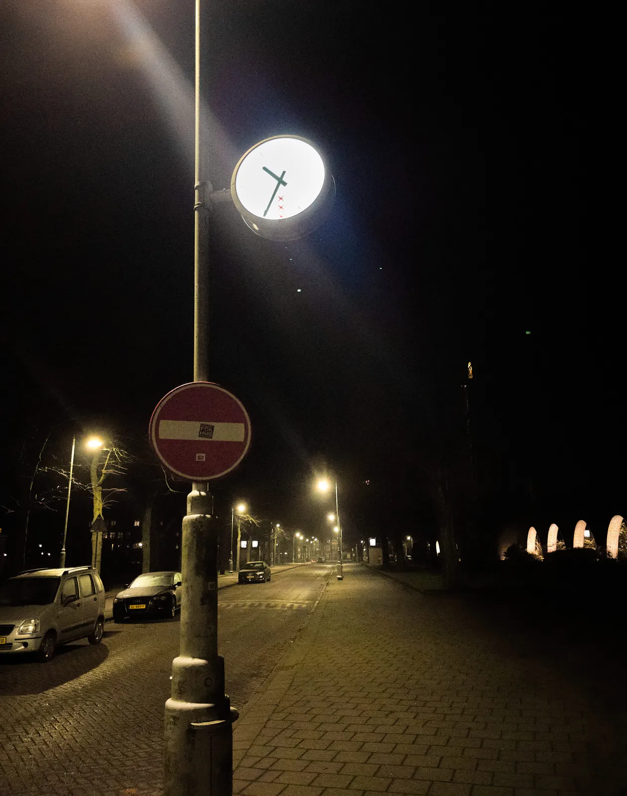 A very dark night, with some street lights. A few cars are parked on the left, and on the right there are some dimly lit arches. There’s a clock on a pole in the center of the image. It points at 35 minutes past 10.