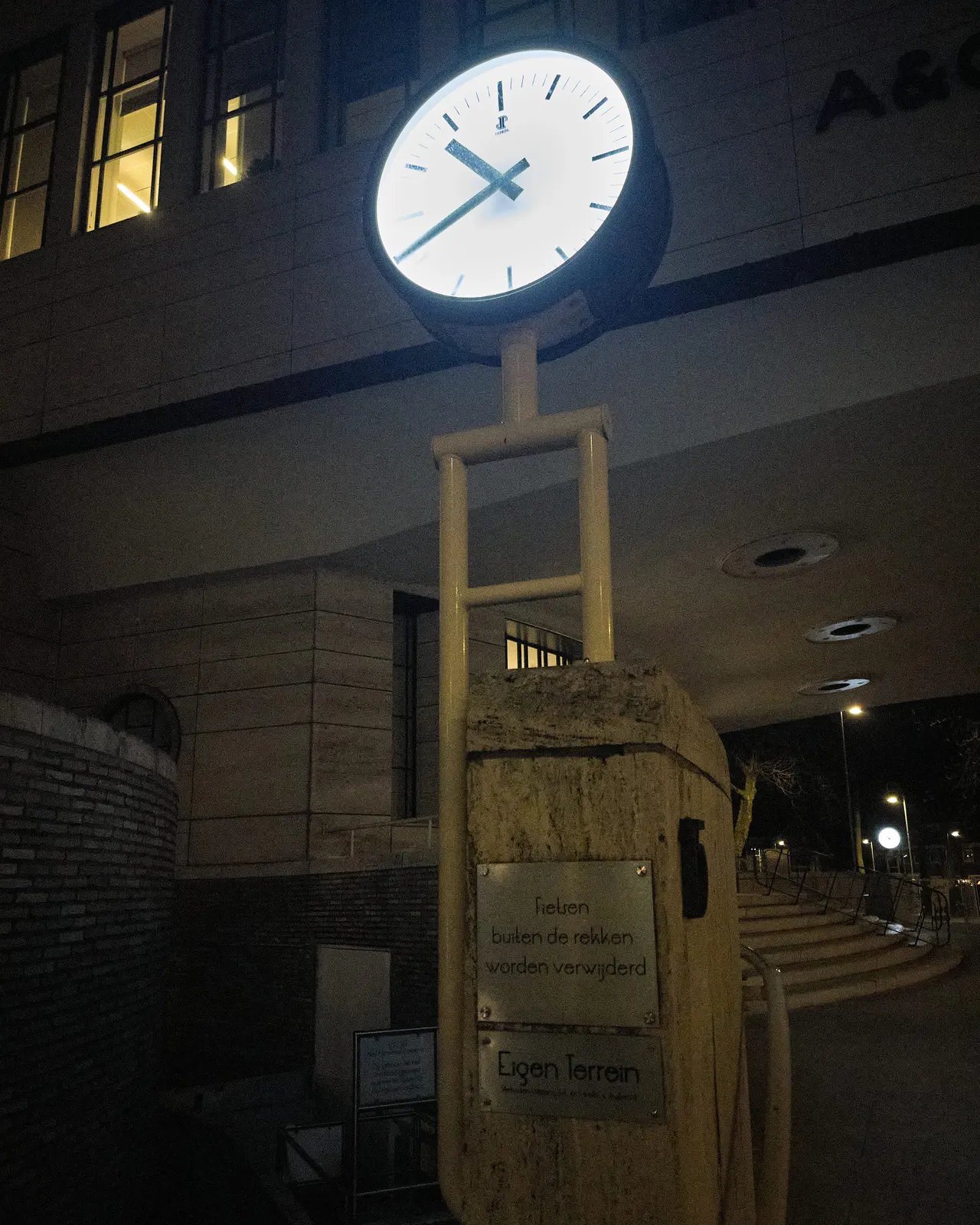 In the background there’s a large office building and in the foreground there’s a brightly lit clock. It points at 18 minutes to 11.