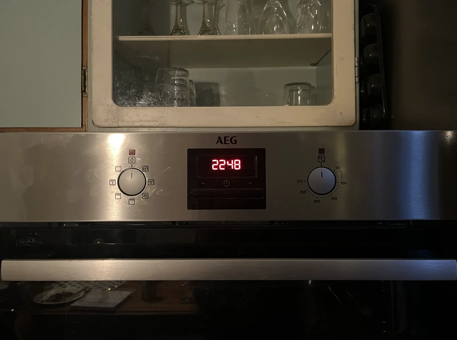 An oven with two knobs and a digital display. Above the oven is a cupboard with glasses. On the digital display the time is shown as 22:48