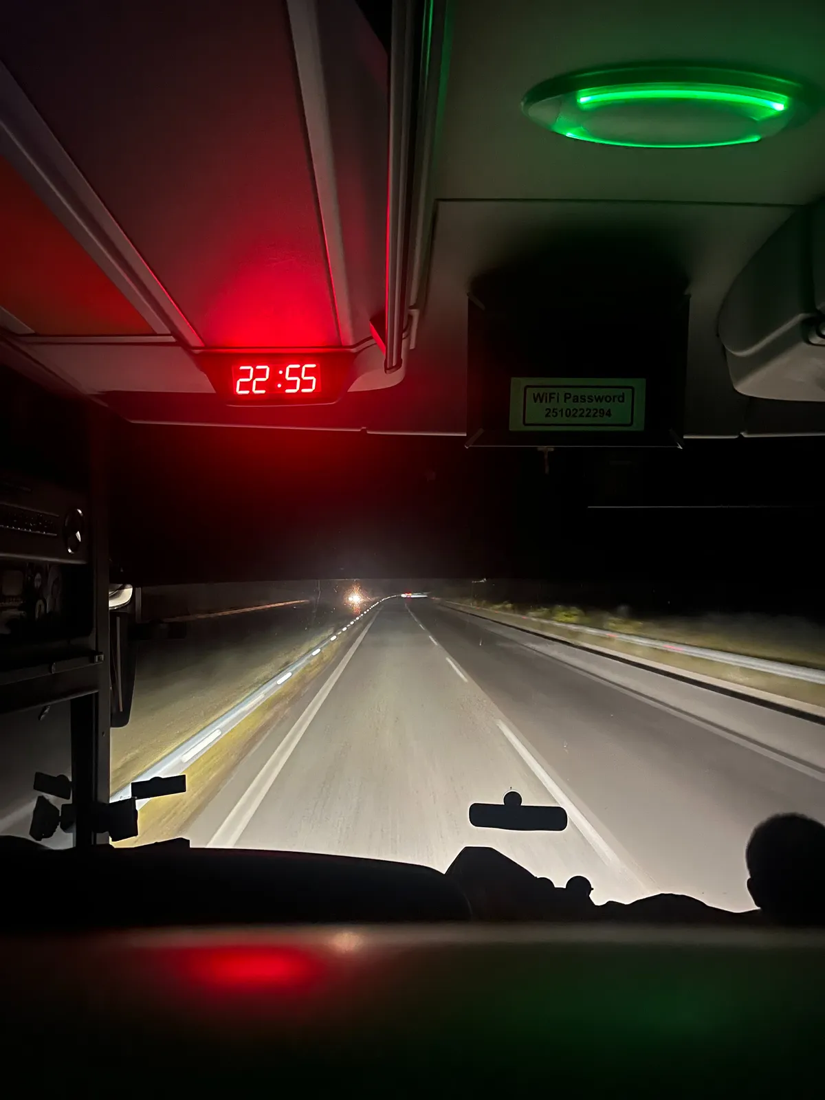 Looking through the front window of a bus, at the dark highway. There’s a digital clock with red digits which says the time is 22:55