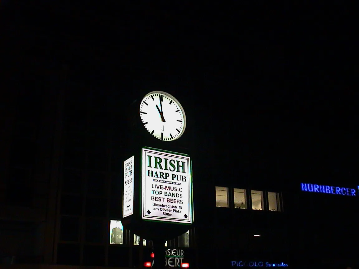 It is one minute to eleven on this big clock above a sign for an Irish Harp Pub.