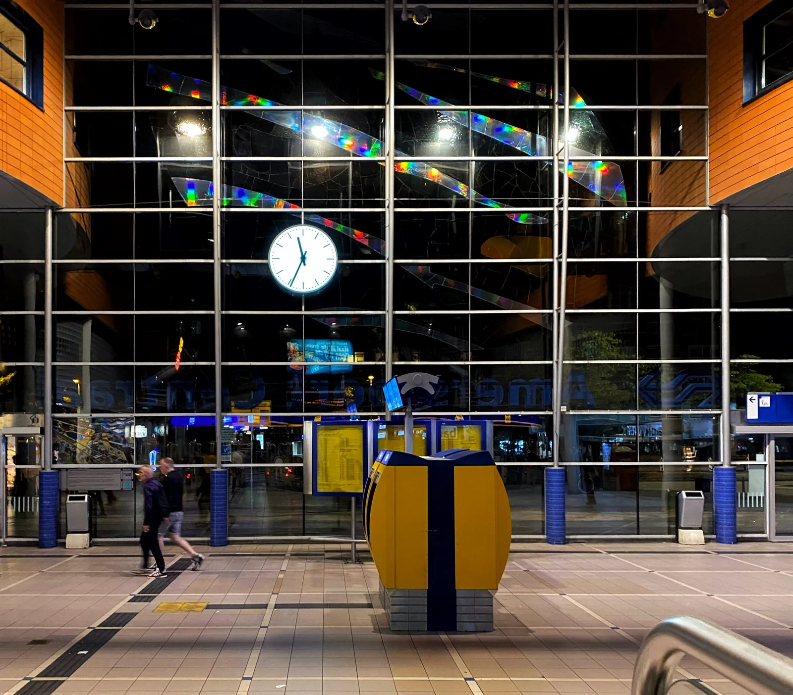 Looking through a high glass facade to the darkness outside. Inside is the lit interior of a Dutch train station. Two travellers stroll by in the background, past a ticket vending machine. In the glass facade there’s a clock that points at 34 minutes past 11.