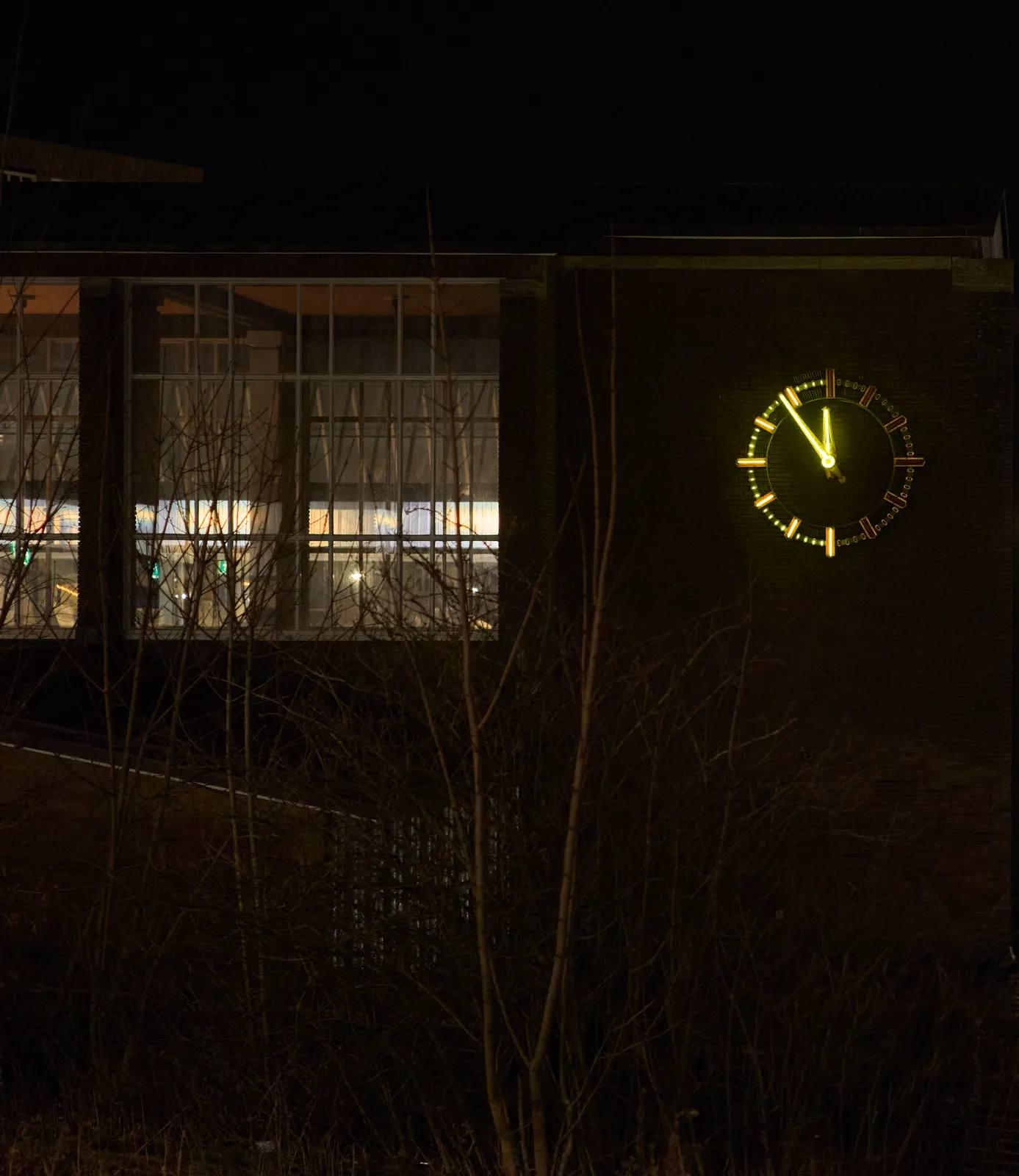 A scene at night. There’s a large clock attached to an old brick building. Half the indicators of the clock (the ones from 30 to 59 minutes) are lit, the other half is not. The hands of the clock are lit as well and the point at 6 minutes to twelve.