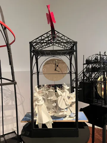 A work of art made from pieces of paper. It’s a scene of people with musical instruments, in an open tent-like structure, with a red bird on top. It also has a large white clock in it. The clock points at one o’clock.
