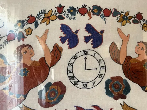 A detail of a piece of embroidery. There are two angels, two birds, and many flowers on it, surrounding a clock that points at three o’clock