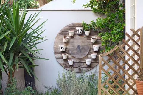 On a wall in a garden, surrounded by plants, is a large clock made from flowering pots. This clock points at 26 minutes past 3