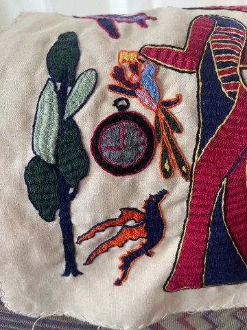A detail of an embroidery. There’s an embroidered clock on it which points at nine o’clock. 